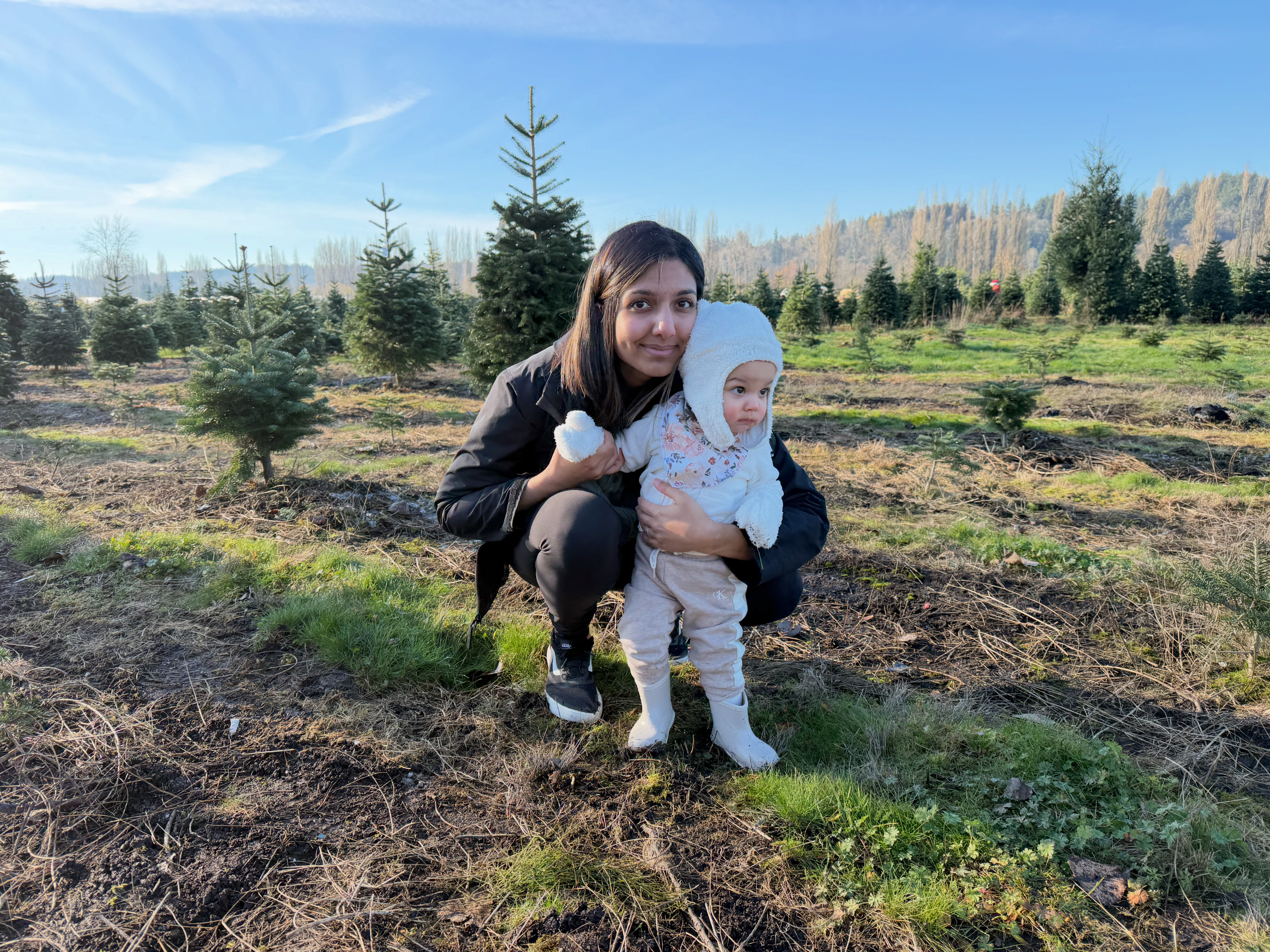 mom crouched in a field of pine trees with her arms around her young daughter